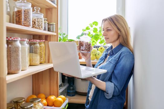 Middle-aged woman in kitchen in pantry with laptop showing cans of food. Female food blogger, online communication, healthy eating video recording, healthy food, technology, hobbies and leisure