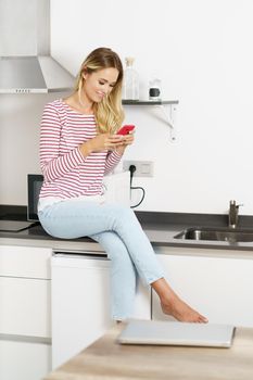 Happy woman using her smartphone sitting in the kitchen at home