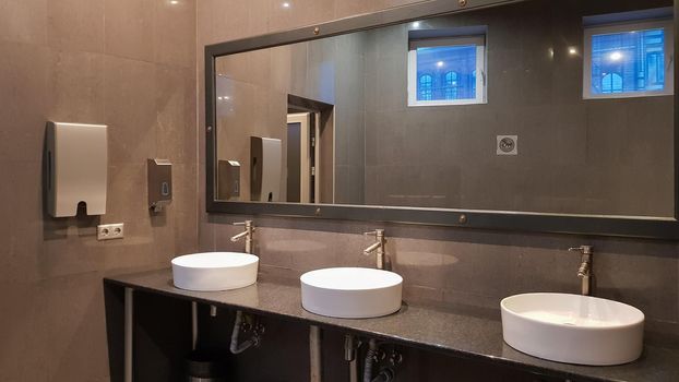 Chrome faucets with white round washbasins in a public toilet with a large mirror and gray walls, modern interior of a public toilet