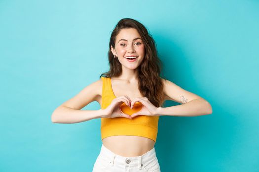 Summer holidays and emotions concept. Young romantic girl showing I love you heart gesture and smiling, standing over blue background. Copy space