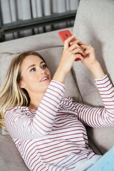 Smiling caucasian woman using her smartphone lying on the sofa at home.