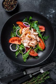 Salad with baked salmon fillet steak, fresh arugula and tomato in a plate. Black background. Top view.