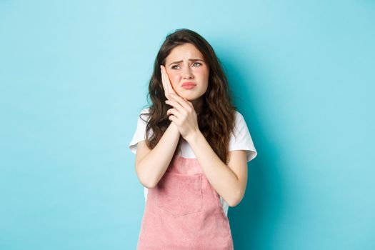 Portrait of frowning girl touching cheek, having toothache, looking at upper left corner stomatology clinic banner, suffering tooth decay, standing against blue background.