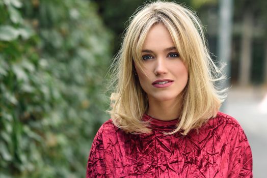 Beautiful blonde woman in green leaves background. Young girl wearing red dress standing in the street. Pretty female with frizzy hairstyle and blue eyes.