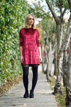 Beautiful blonde woman in urban background. Young girl wearing red dress and tights standing in the street. Pretty female with straight hairstyle and blue eyes.