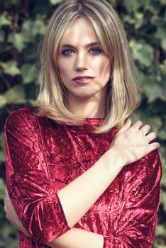 Beautiful blonde woman in green leaves background. Young girl wearing red dress standing in the street. Pretty female with frizzy hairstyle and blue eyes.