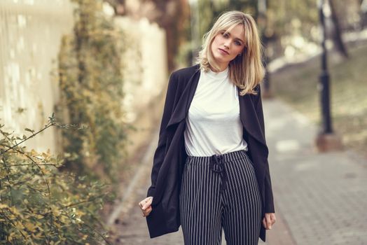 Beautiful blonde woman in urban background. Young girl wearing black blazer jacket and striped trousers standing in the street. Pretty female with straight hair hairstyle and blue eyes.