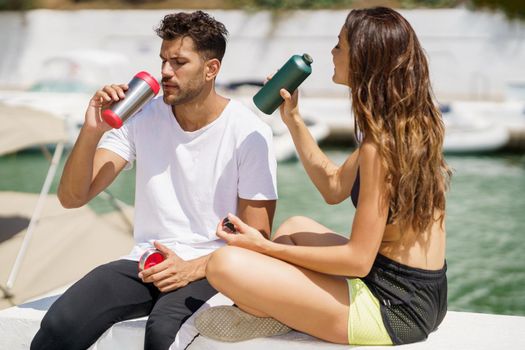 Young fitness people hydrate themselves with water in metal bottles while taking a break after sport.