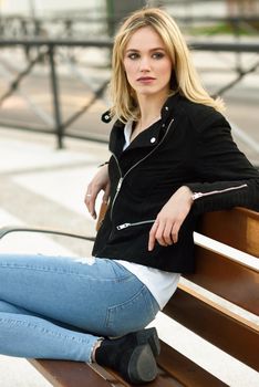 Attractive blonde woman in urban background. Young girl wearing black zipper jacket and blue jeans trousers sitting on a bench in the street. Pretty female with straight hair hairstyle and blue eyes.