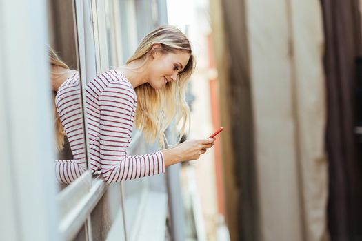 Caucasian woman leaning out of her house window using a smart phone.