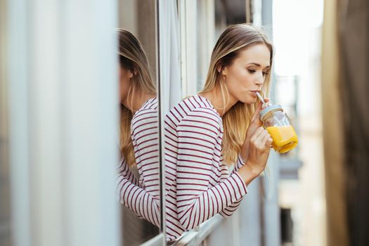 Young caucasian woman drinking a glass of natural orange juice, leaning out the window of her home.
