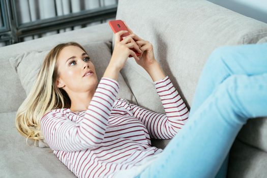 Young caucasian woman using her smartphone lying on the sofa at home.