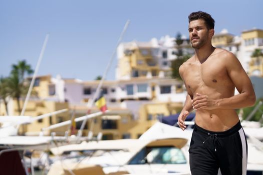 Young man with an athletic body running shirtless through a harbour full of boats.