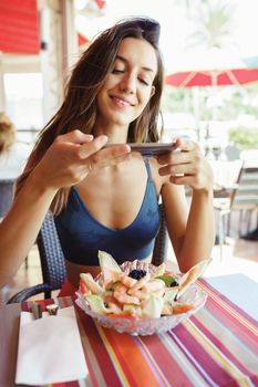 Young woman photographing her salad with a smartphone while sitting in a coastal restaurant