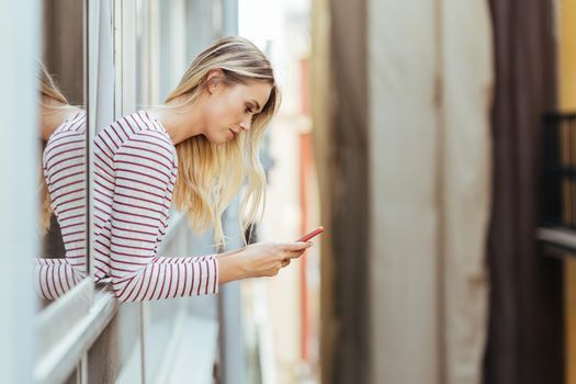 Attractive woman leaning out of her house window using a smart phone.