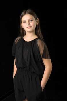 Cute long haired girl posing half turned to camera. Portrait of beautiful serious sad girl dressed in summer black jumpsuit standing against black background in studio