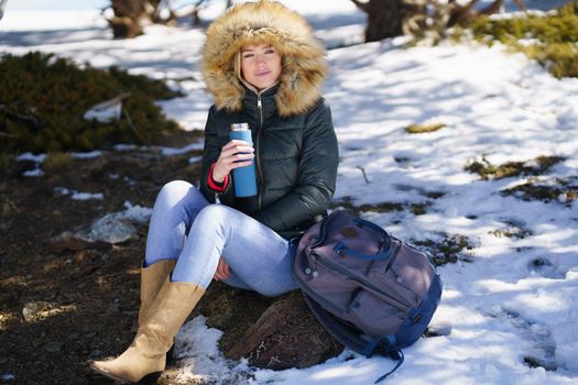 Young woman drinking something hot from a metal thermos bottle sitting on a rock in the snowy mountains., in Sierra Nevada, Granada, Spain. Female wearing winter clothes.