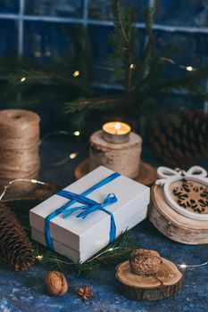 Christmas white gift box with blue bow ribbon, wooden decorations, candle, nuts on magic dark blue background. Vintage Xmas greeting card mockup, postcard