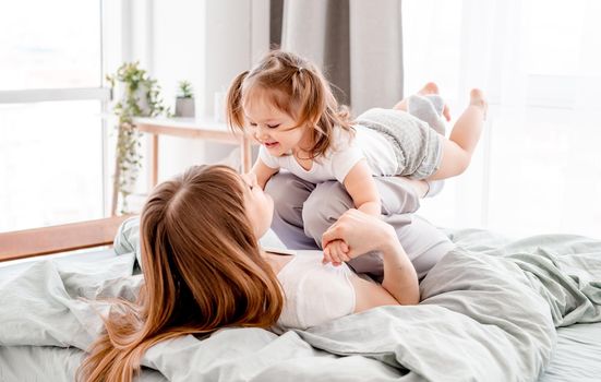 Young mother lying in the bed and swinging cute daughter on her legs. Caucasian mom and her child have fun in the bedroom with sunlight. Beautiful family morning time together