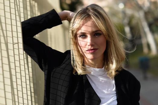 Beautiful blonde woman in urban background. Young girl wearing black blazer jacket standing in the street. Pretty female with straight hair hairstyle and blue eyes.