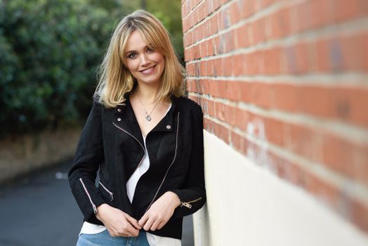 Attractive blonde woman smiling in urban background. Young girl wearing black zipper jacket and blue jeans trousers standing in the street. Pretty female with straight hair hairstyle and blue eyes on brick wall.