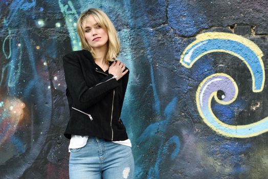 Attractive blonde woman in urban background. Young girl wearing black zipper jacket and blue jeans trousers standing in the street. Pretty female with straight hair hairstyle and blue eyes.