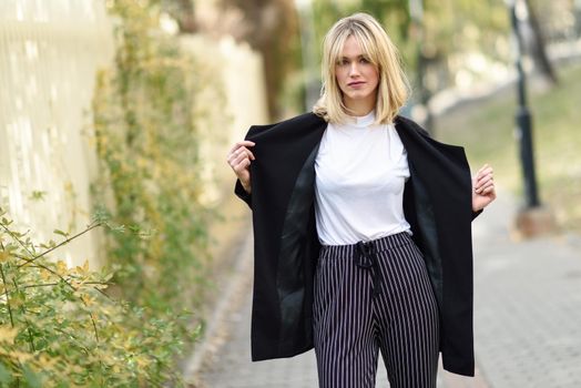 Beautiful blonde woman in urban background. Young girl wearing black blazer jacket and striped trousers standing in the street. Pretty female with straight hair hairstyle and blue eyes.