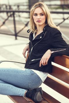 Attractive blonde woman in urban background. Young girl wearing black zipper jacket and blue jeans trousers sitting on a bench in the street. Pretty female with straight hair hairstyle and blue eyes.