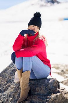 Young blonde woman sitting on a rock in the snowy mountains in winter, in Sierra Nevada, Granada, Spain. Female wearing winter clothes.