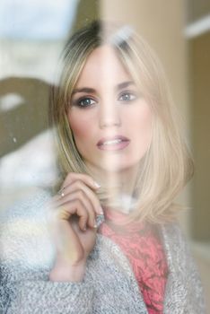 Thoughtful young blonde woman with lost eyes from the window. Beautiful girl at home wearing winter clothes. Pretty female with straight hair and blue eyes.