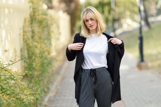 Beautiful blonde woman in urban background. Young girl wearing black blazer jacket and striped trousers standing in the street. Pretty female with straight hair hairstyle and blue eyes.