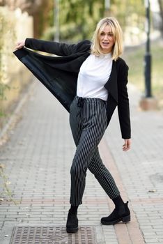 Funny blonde woman smiling in urban background. Young girl wearing black blazer jacket and striped trousers standing in the street. Pretty female with straight hair hairstyle and blue eyes.