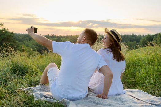 Beautiful adult couple together taking selfie photo on smartphone. Happy man and woman sitting on grass outdoors in sunset sunlight. Relationships, love, date, people 30s 40s age