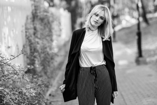 Beautiful blonde woman in urban background. Young girl wearing black blazer jacket and striped trousers standing in the street. Pretty female with straight hair hairstyle and blue eyes.