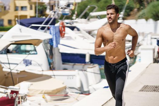 Young man with an athletic body running shirtless through a harbour full of boats.