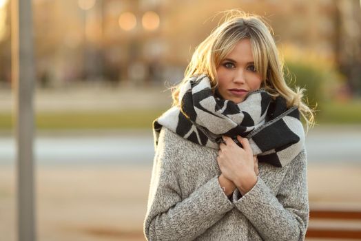 Attractive blonde woman in urban background with sun backlight. Young girl wearing winter coat and scarf standing in the street. Pretty female with straight hair hairstyle and blue eyes.