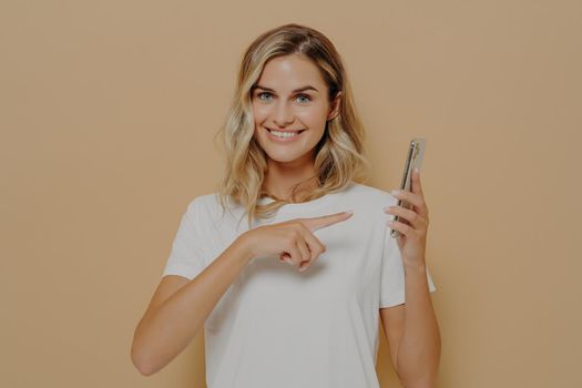 Cute excited female pointing on smartphone with forefinger and looking at camera with broad smile, reading good news in internet and feeling happy while posing isolated over beige background