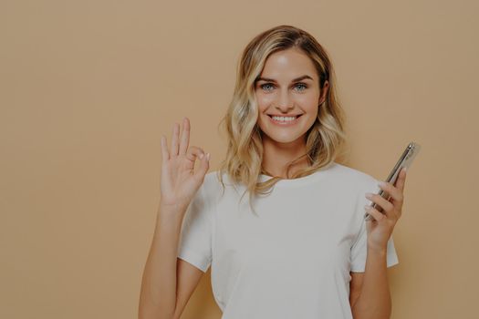 Happy cheerful female in casual tshirt holding smartphone and showing okay gesture, being happy after pleasant conversation or hearing good news, isolated over beige background