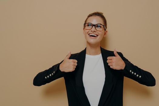 Portrait of positive female customer in eyeglasses with broad smile showing thumbs up with both hands and looking at copy space with happy expression while posing isolated over beige background