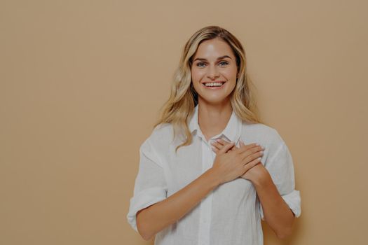 Portrait of admiring cute woman in love with blond hair holding hands on chest sighing with romantic smile, gazing at camera as feeling grateful and appreciative isolated on beige background
