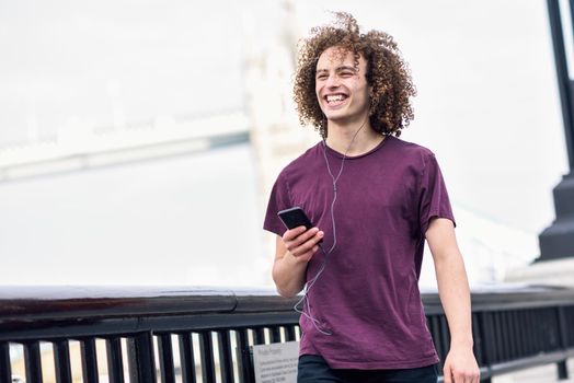 Young man listening to the music with earphones and smart phon in urban background. Thames River in London.