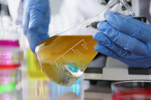 Close-up of scientist in protective gloves holding flask with yellow liquid in medical chemistry lab. Chemist doing experiment in laboratory. Science, discovery, test concept