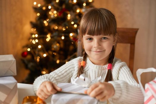 Girl packing present box while sitting at table in living room, looks at camera with charming smile, having pigtails, wearing white sweater, posing with xmas tree on background.