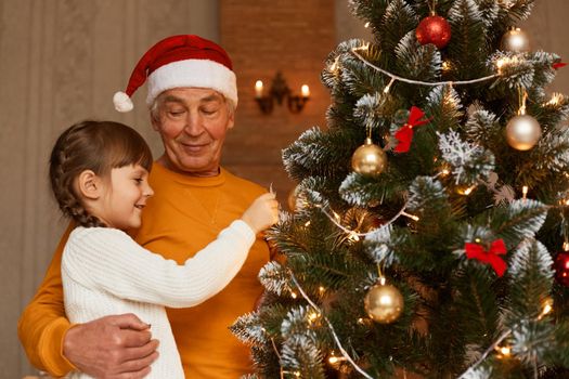 Indoor shot of happy mature man in santa hat and orange sweater posing near x mas tree with his cute charming granddaughter in festive room at home, merry christmas.