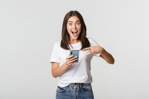 Excited brunette girl smiling amazed and pointing at smartphone screen.