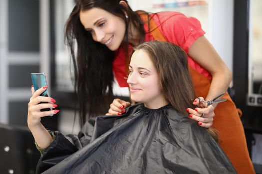 Portrait of smiling client on appointment at hairdresser. Woman show internet content to barber girl on smartphone. Change image, enter new life. Barbershop, style, beauty salon concept