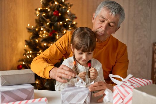 Mature man posing in festive living room with kid, packing Christmas gifts near fir tree, looking concentrated at boxes, child in white casual shirt, mature male in orange jumper.