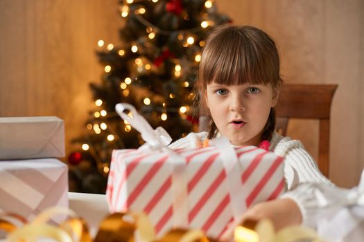 Astonished girl receiving gift from Santa Claus, looking at present box with big eyes, looks surprised and shocked, child sitting at table with xmas tree on background.
