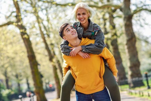 Funny couple in a urban park. Boyfriend carrying his girlfriend on piggyback. Love and tenderness, dating, romance. Lifestyle concept