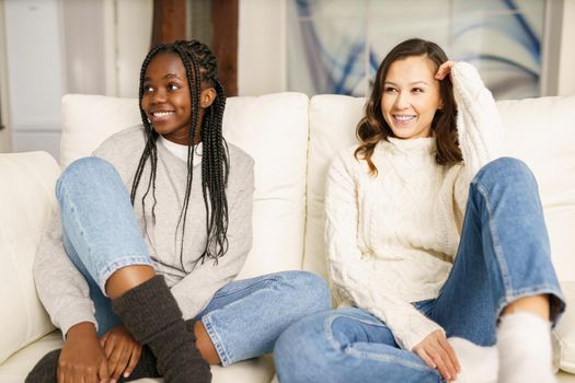 Two female student friends smiling together on the couch at home. Multiethnic women.